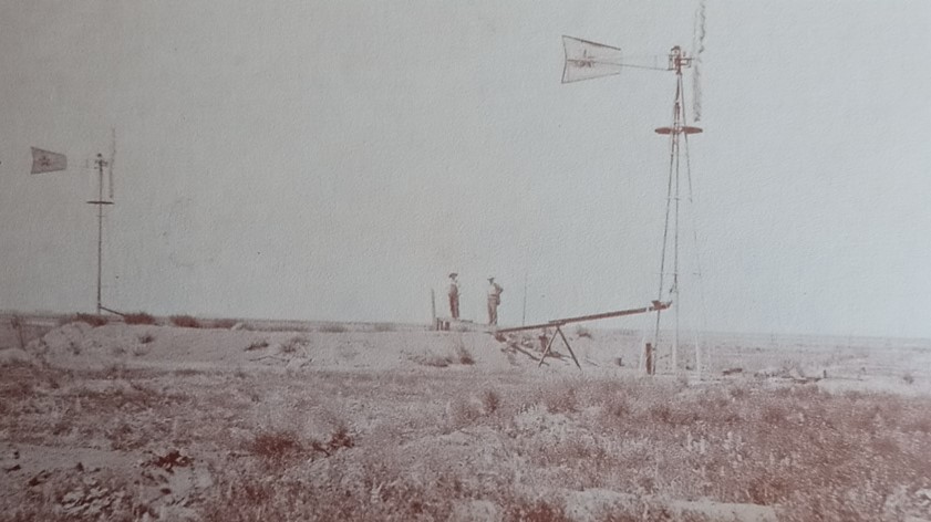 Photo of two men standing near two windmills on a farm.